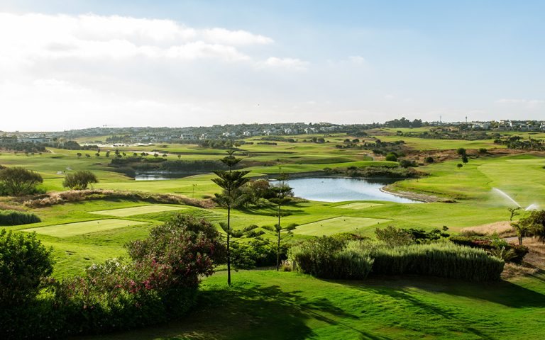 Vue d'ensemble de Palm Casablanca : fairways vallonnés, pièce d'eau bordée de roseaux, arbres de haute tige et ville au loin.