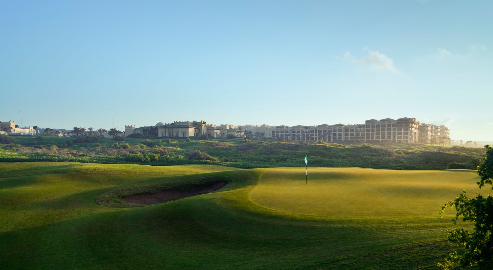 Green de Mazagan vu du départ : modelés en creux et en butte au premier plan, bâtiments du resort alignés sur la crête en arrière-plan.