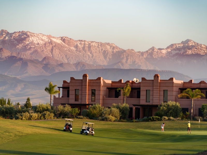 Le clubhouse de Palm Ourika en terre ocre, joueurs et voiturettes sur le fairway, sommets enneigés du Haut Atlas en arrière-plan.